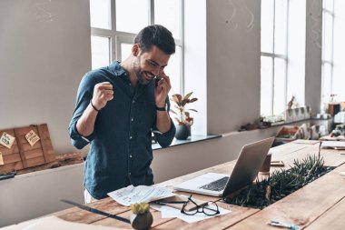 mutlu yakışıklı genç adam akıllı telefon duran tahta masada modern ofis içinde laptop ile konuşurken