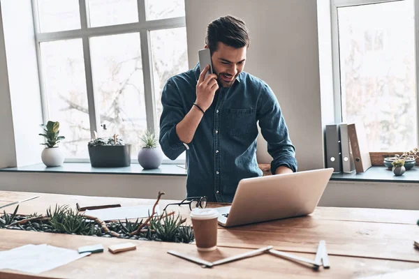 近代的なオフィスにラップトップと木製のテーブルで立ちながらスマート フォンで話して幸せのハンサムな若い男 — ストック写真