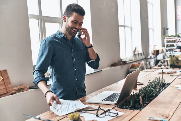 近代的なオフィスにラップトップと木製のテーブルで立ちながらスマート フォンで話して幸せのハンサムな若い男 — ストック写真