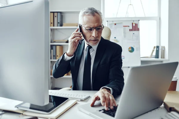 Homme âgé en costume d'affaires élégant parlant au téléphone tout en travaillant dans un bureau moderne — Photo