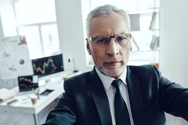 Zelfportret van senior man in elegant zakenpak kijkend naar de camera terwijl hij op kantoor staat — Stockfoto
