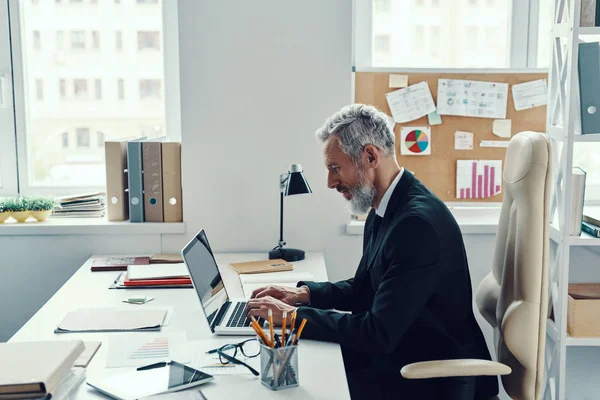 Homme mûr concentré en costume complet en utilisant un ordinateur portable tout en travaillant dans un bureau moderne — Photo