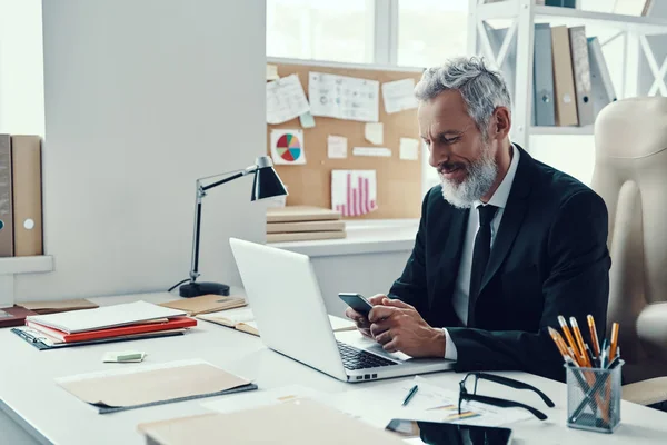 Homme mûr réfléchi en costume complet souriant et utilisant un téléphone intelligent tout en travaillant dans un bureau moderne — Photo