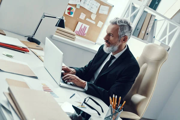 Homme mûr concentré en costume complet en utilisant un ordinateur portable tout en travaillant dans un bureau moderne — Photo