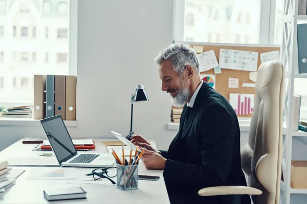 Συμπυκνωμένο ώριμο άτομο σε πλήρη κοστούμι χρησιμοποιώντας ψηφιακή ταμπλέτα, ενώ εργάζονται στο σύγχρονο γραφείο — Φωτογραφία Αρχείου