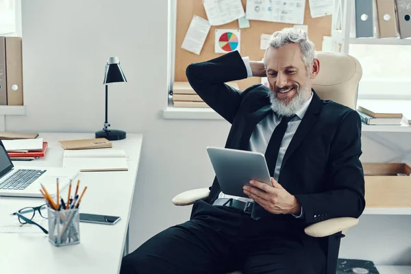 Heureux homme mature en costume complet à l'aide d'une tablette numérique et souriant tout en travaillant dans un bureau moderne — Photo