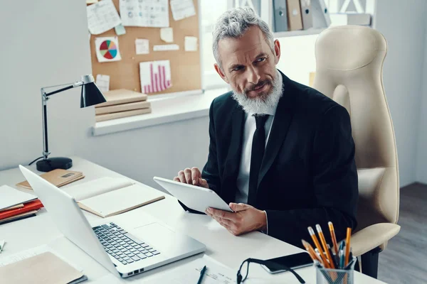 Thoughtful mature man in full suit using digital tablet and looking away while working in modern office — Stock Photo, Image