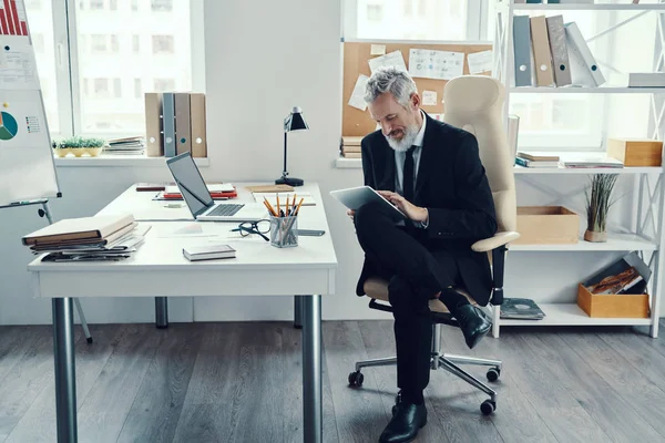 Thoughtful mature man in full suit using digital tablet while working in modern office — Stock Photo, Image