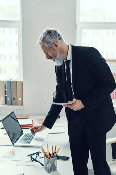 Προσεκτική ώριμος άνθρωπος σε πλήρη κοστούμι χρησιμοποιώντας φορητό υπολογιστή και ψηφιακό tablet, ενώ εργάζονται στο σύγχρονο γραφείο — Φωτογραφία Αρχείου