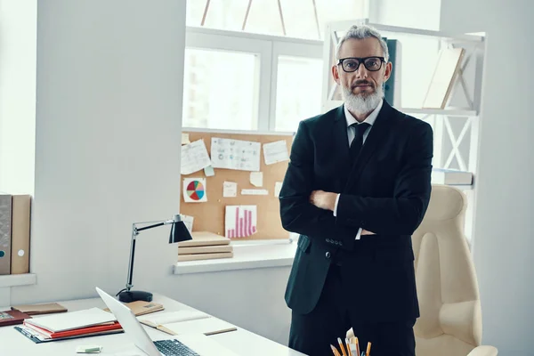 Confident mature man in full suit looking at camera and keeping arms crossed while working in modern office — Stock Photo, Image