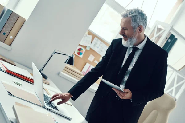 Задумчивый зрелый мужчина в полном костюме с помощью ноутбука и цифрового планшета во время работы в современном офисе — стоковое фото