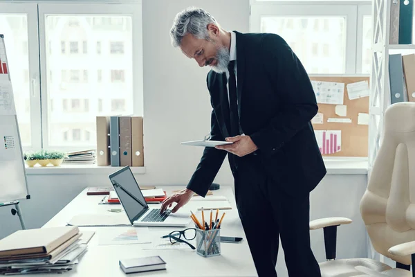 Homme mûr réfléchi en costume complet en utilisant un ordinateur portable et une tablette numérique tout en travaillant dans un bureau moderne — Photo