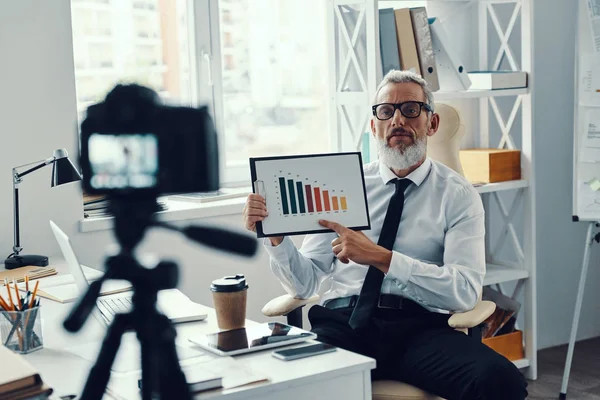 Confident mature man in elegant shirt and tie showing chart while making social media video — Stock Photo, Image
