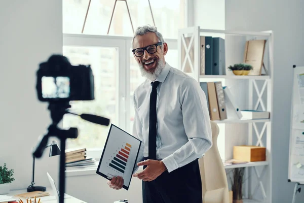 Happy mature man in elegant shirt and tie showing chart and sharing business experience while making social media video — Stock Photo, Image