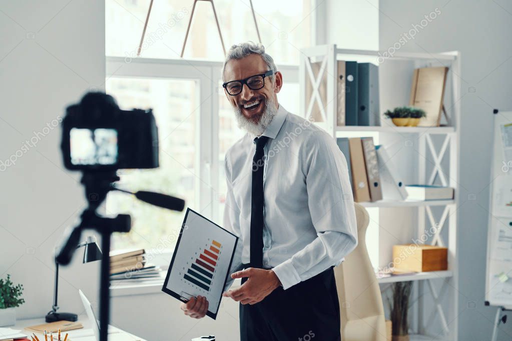 Happy mature man in elegant shirt and tie showing chart and sharing business experience while making social media video