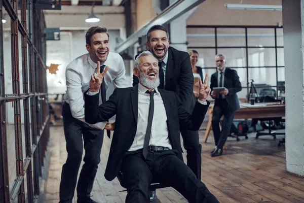 Vidám Üzletemberek Tolják Főnöküket Irodai Székre Miközben Futnak Folyosón — Stock Fotó