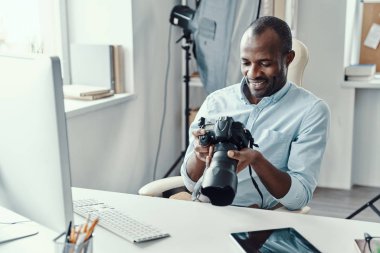 Yakışıklı genç Afrikalı adam dijital kamera tutuyor ve modern ofiste çalışırken gülümsüyor.     