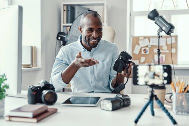 Çekici genç Afrikalı gömlekli adam dijital kamera gösteriyor ve sosyal medya videosu çekerken bir şeyler anlatıyor.      