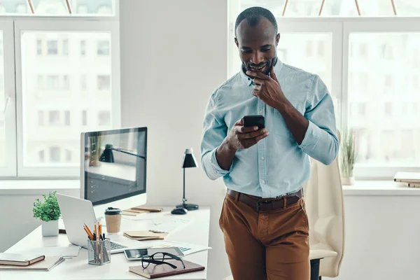 オフィスで働いている間 スマートフォンと笑顔を使用してシャツに身を包んだ若いアフリカ人男性を自信を持って — ストック写真