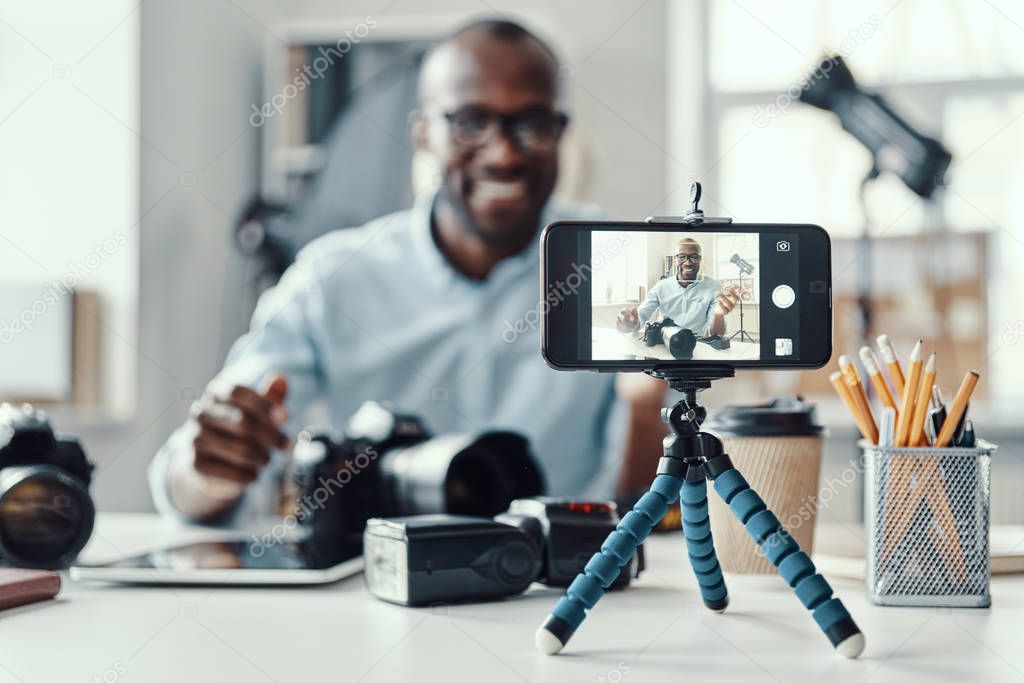 Happy young African man in shirt showing digital camera and telling something while making social media video
