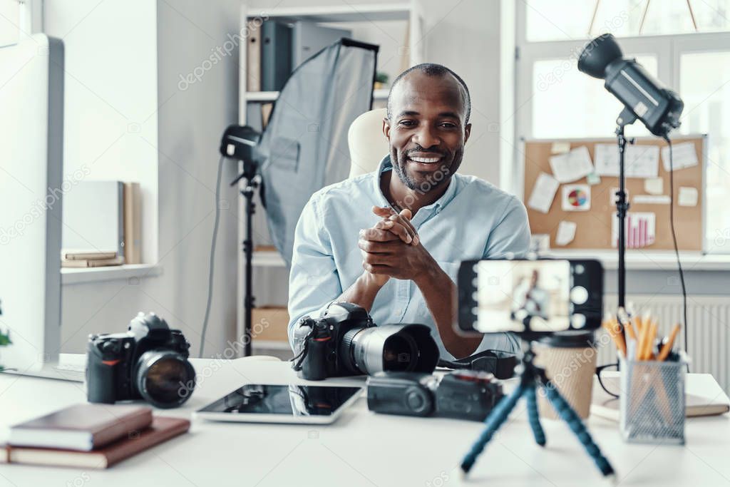 Charming young African man in shirt showing digital camera and telling something while making social media video