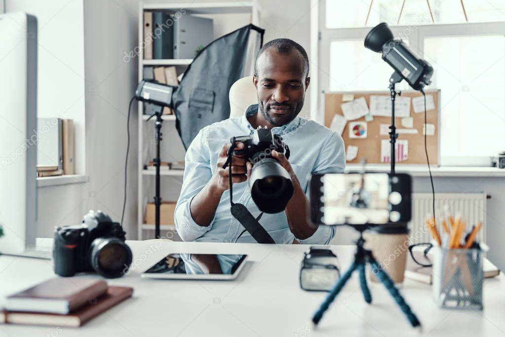 Charming young African man in shirt showing digital camera and telling something while making social media video