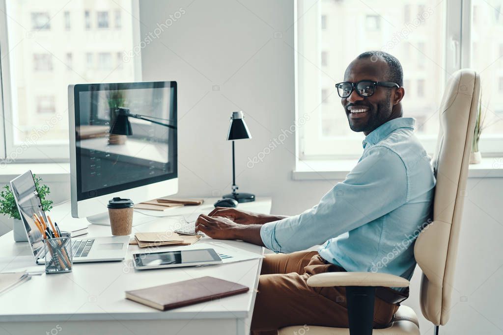 Happy young African man in shirt using computer and smiling while working in the office   
