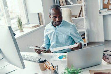 Ofiste çalışırken, tişörtlü genç Afrikalı bir adamın satış verilerini analiz etmesinin en iyi görüntüsü.