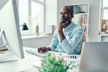 Ofiste çalışırken bilgisayar kullanan tişörtlü düşünceli bir Afrikalı.           