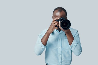 Çekici genç Afrikalı adam gri arka planda dikilirken dijital kamera kullanarak fotoğraf çekiyor.        