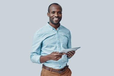 Dijital tablet kullanan ve gri arka planda dururken gülümseyen mutlu genç Afrikalı adam.              