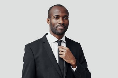 Resmi giyinmiş yakışıklı Afrikalı genç adam kameraya bakıyor ve gri arka planda dururken kravatını düzeltiyor.     