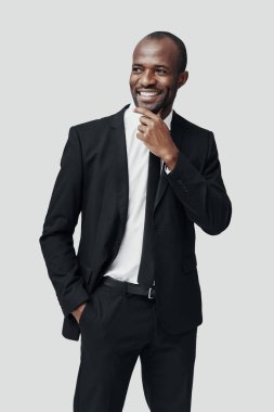 Resmi giyinmiş şık genç Afrikalı adam gri arka planda dururken başka tarafa bakıyor ve gülümsüyor.      