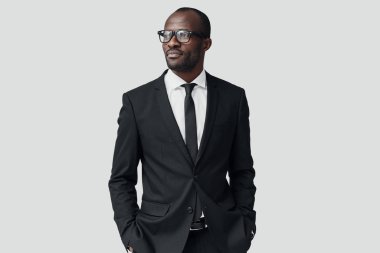 Resmi giyinmiş kendinden emin genç Afrikalı adam gri arka planda dikilirken başka tarafa bakıyor.