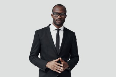 Resmi giyinmiş kendinden emin genç Afrikalı adam kameraya bakıyor ve gri arka planda dururken ellerini sıkıca tutuyor.