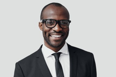 Resmi giyinmiş kendinden emin genç Afrikalı adam gri arka planda dururken kameraya bakıyor ve gülümsüyor.