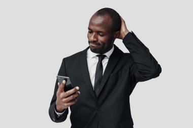 Resmi giyinmiş düşünceli genç Afrikalı adam gri arka planda dururken akıllı telefon kullanıyor.         