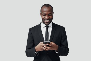 Resmi giyinmiş modern Afrikalı genç adam gri arka planda dururken akıllı telefon kullanıyor ve gülümsüyor.                 