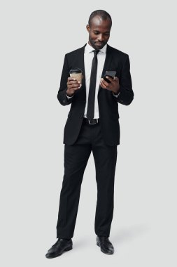 Gri arka planda dururken akıllı telefon kullanan ve gülümseyen resmi giyimli modern Afrikalı genç adam.