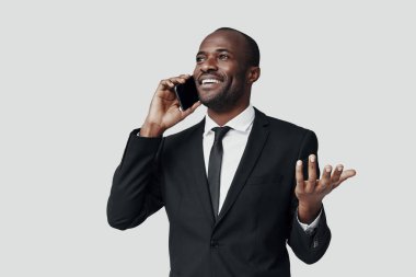 Resmi kıyafetli mutlu Afrikalı bir genç, gri arka planda dururken telefonla konuşuyor ve gülümsüyor.