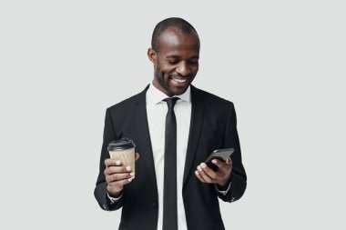 Resmi giyinmiş modern Afrikalı genç adam gri arka planda dururken akıllı telefon kullanıyor ve gülümsüyor.