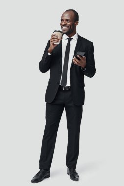 Resmi giyinmiş, akıllı telefon kullanan ve gri arka planda dururken gülümseyen tam boy modern Afrikalı adam.   