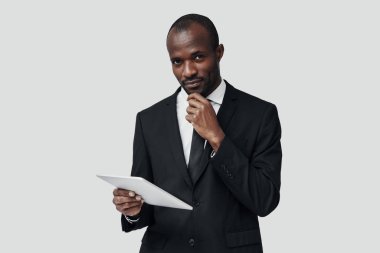 Gri arkaplana karşı ayakta dururken dijital tablet kullanarak resmi olarak çalışan başarılı genç Afrikalı adam    