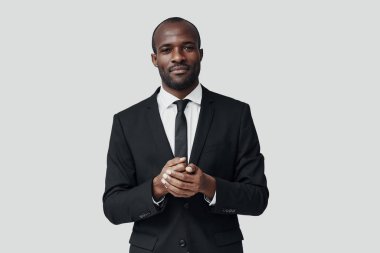 Resmi giyinmiş yakışıklı Afrikalı genç adam gri arka planda dururken kameraya bakıyor ve gülümsüyor. 