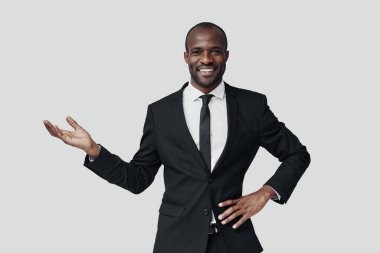 Resmi giyinmiş yakışıklı Afrikalı genç adam fotokopi alanını işaret ediyor ve gri arka planda dururken gülümsüyor.