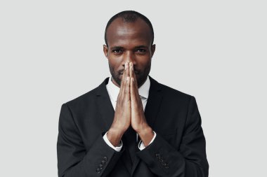 Resmi giyinmiş genç Afrikalı adam gri arka planda dururken ellerini sıkıca tutuyor.   