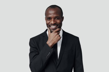 Resmi giyinmiş şık genç Afrikalı adam gri arka planda dururken kameraya bakıp gülümsüyor.