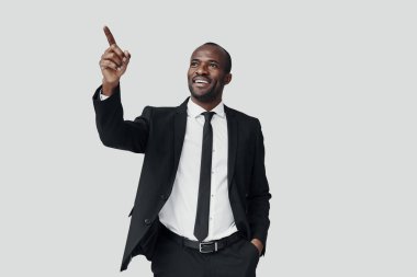 Resmi giyinmiş yakışıklı Afrikalı genç adam fotokopi alanını işaret ediyor ve gri arka planda dururken gülümsüyor.