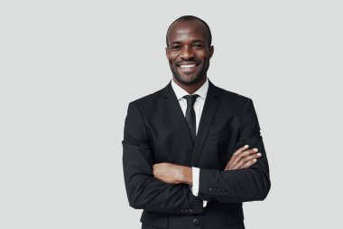 Resmi giyinmiş yakışıklı Afrikalı genç adam gri arka planda dururken kameraya bakıyor ve gülümsüyor.                       