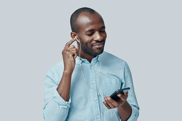 迷人的非洲年轻人一边在灰色背景下一边用智能手机听音乐 一边微笑 — 图库照片